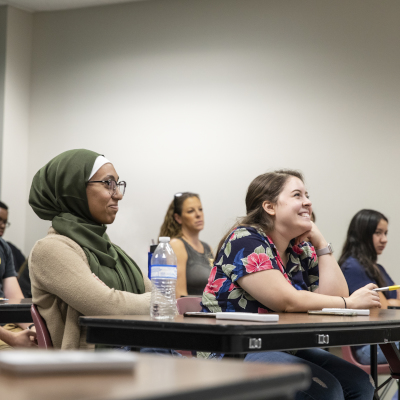 A classroom setting with students attentively listening to a lecture. The foreground features a student wearing a green hijab and glasses, smiling as she engages in the lesson. Other students are visible in the background, contributing to the atmosphere of focused learning.
