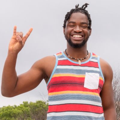 |奥斯汀社区学院区 A smiling young man wearing a colorful striped tank top, standing outdoors and making the “Hook ’em Horns” hand sign, a symbol associated with the University of Texas at Austin. The image conveys pride and enthusiasm for the 速度 co-enrollment program with UT Austin.