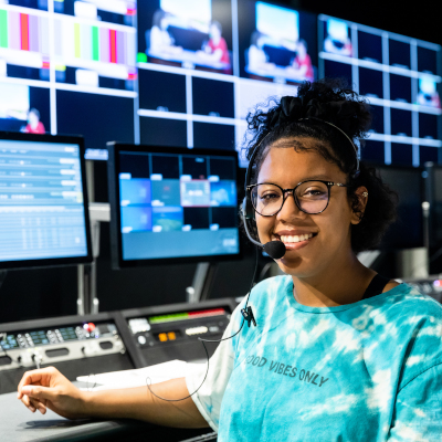 高地校区|奥斯汀社区学院区 A young woman with glasses and a headset, smiling while sitting in front of a control panel in a media production room. Multiple screens showing various video feeds are visible in the background.