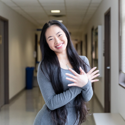 A smiling young woman with long black hair stands in a hallway with her arms crossed. She exudes confidence and warmth, representing the welcoming environment of the AAPI center.