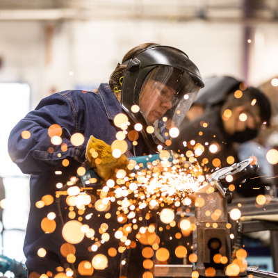 埃尔金校区|奥斯汀社区学院区 A student wearing protective gear, including a face shield and gloves, is using a welding tool, with bright sparks flying in the foreground. The student is focused on their work in a workshop environment.