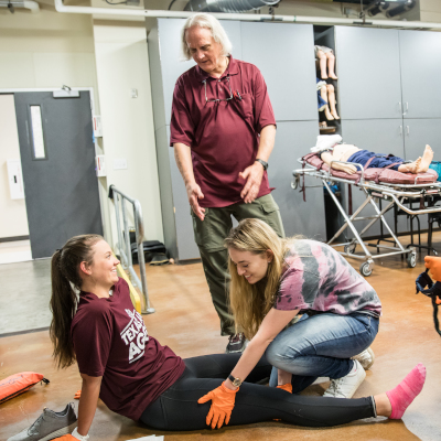 柏树溪b|奥斯汀社区学院区 A scene of an EMT training session where a female student practices emergency care on another student lying on the floor, while an instructor observes.