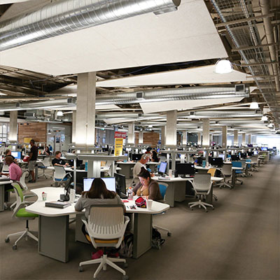高地校区|奥斯汀社区学院区 A large, open workspace filled with students sitting at desks with computers, working individually and in small groups. The room has modern lighting and exposed ductwork, giving it a contemporary, industrial feel.