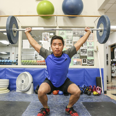  A determined student lifting a barbell with weights in a fitness center. The background shows gym equipment, including kettlebells and exercise balls, emphasizing the active environment.